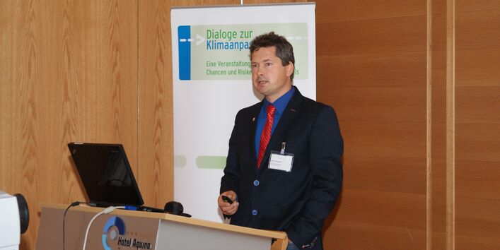 Vortragener steht am Rednerpult. Ein Laptop mit geöffnetem Deckel steht auf dem Rednerpult. Im Hintergrund steht der Veranstaltungsaufsteller Dialoge zur Klimaanpassung.