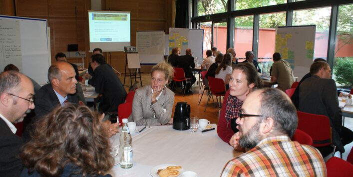 Teilnehmer sitzen an Gruppenarbeitstischen auf roten Stühlen im Veranstaltungsraum vor einer großen Fensterfront. Im Hintergrund stehen Moderationswände.