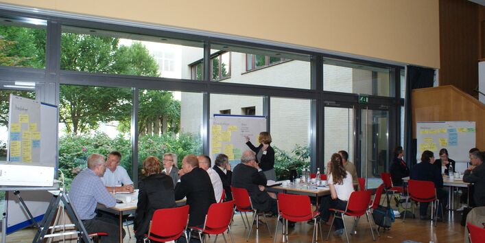 Teilnehmer sitzen an Gruppenarbeitstischen im Raum vor einer großen Fensterfront. Eine Teilnehmerin schreibt im Hintergrund Text auf Papier an Moderationswand.