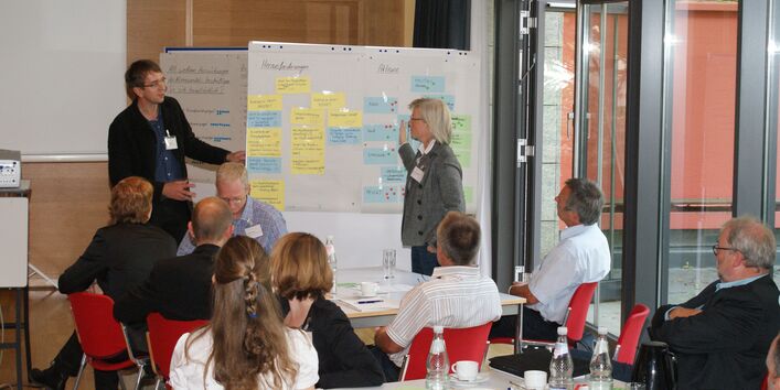 Zwei Teilnehmer stehen an Moderationswand im Dialog miteinander. An der Moderationswand hängen farbige Moderationskarten. Auf der rechten Seite steht die Tür von der Fensterfront offen. Zuhörer sitzen im Vordergrund an Gruppenarbeitstischen.