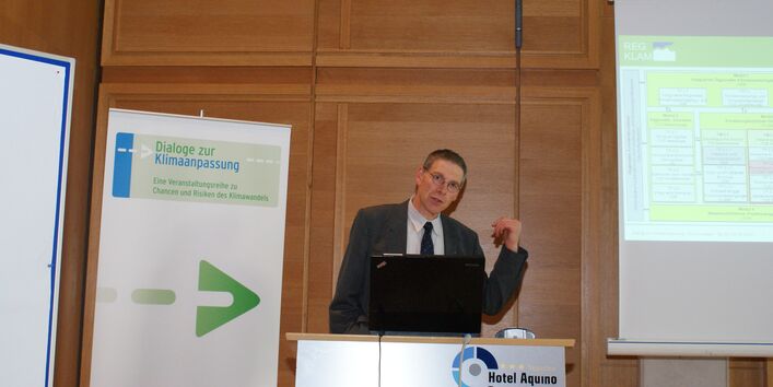 Vortragender steht am Rednerpult und zeigt mit erhobenen Daumen hinter sich auf seine Präsentation, die im Hintergrund auf einer Leinwand zu sehen ist. Im Hintergrund steht der Veranstaltungsaufsteller Dialoge zur Klimaanpassung. 