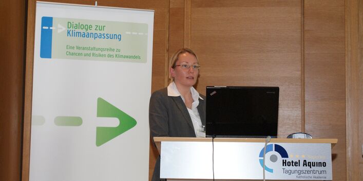 Vortragende steht am Rednerpult vor mit Holz vertäfelter Wand. Im Vordergrund steht am Rednerpult der Schriftzug des Veranstaltungsortes Tagungszentrum Hotel Aquino. Im Hintergrund steht der Veranstaltungsaufsteller Dialoge zur Klimaanpassung.