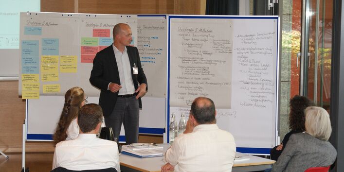 Vortragender steht vor Moderationswänden und präsentiert Ergebnisse. Im Vordergrund sitzen die Zuhörer am Gruppenarbeitstisch.