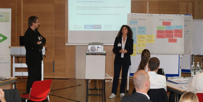 Vortragende präsentieren Ergebnisse. Im Vordergrund sitzen Zuhörer. Im Hintergrund stehen Moderationswände mit farbigen Karten und eine Leinwand. Auf der Leinwand ist eine Präsentationseite Diskussionsfragen Weltcafé 2 zu sehen.
