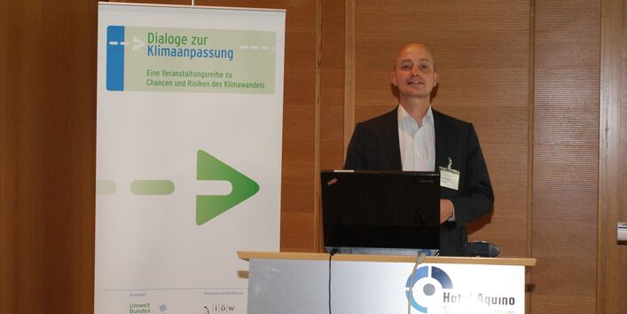 Vortragender steht am Rednerpult vor mit Holz vertäfelter Wand. Das Rednerpult trägt vorne den Schriftzug Hotel Aquino Tagungszentrum. Im Hintergrund steht der Veranstaltungsaufsteller Dialoge zur Klimaanpassung.