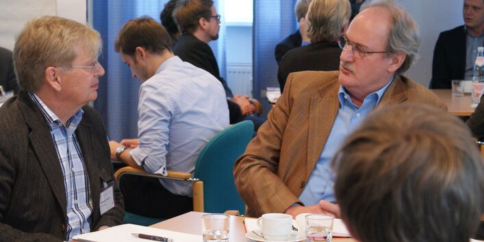 Teilnehmer sitzen am Gruppenarbeitstisch und sprechen miteinander. Im Hintergrund sitzen weitere Teilnehmer an Gruppenarbeitstischen.