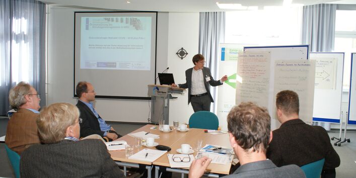 Teilnehmer sitzen am Arbeitgruppentisch und hören der Vortragenden zu. Die Vortragende spricht zu den Teilnehmenden und zeigt auf Themen an Moderationswänden.