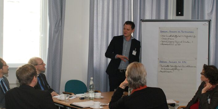 Teilnehmer sitzen am Gruppenarbeitstisch und hören Vortragenden zu. Der Vortragender steht neben einer Moderationswand.