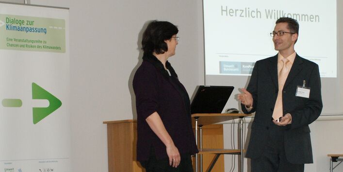 Ein Teilnehmer und eine Teilnehmerin befinden sich im Dialog miteinander. Im Hintergrund sieht man den offenen Laptop auf dem Rednerpult sowie den Veranstaltungsaufsteller und ein Herzliches Willkommen projiziert auf der weißen Wand.