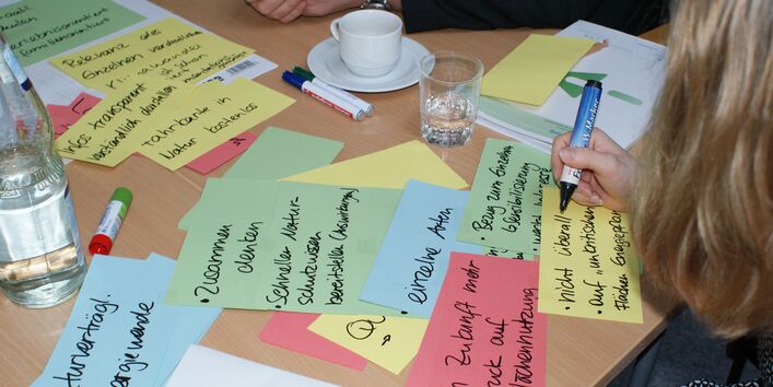 Farbige Moderationskarten liegen beschriftet auf dem Gruppenarbeitstisch. Eine Teilnehmerin beschreibt eine gelbe Moderationskarte zum Thema Naturschutz durch Anpassung