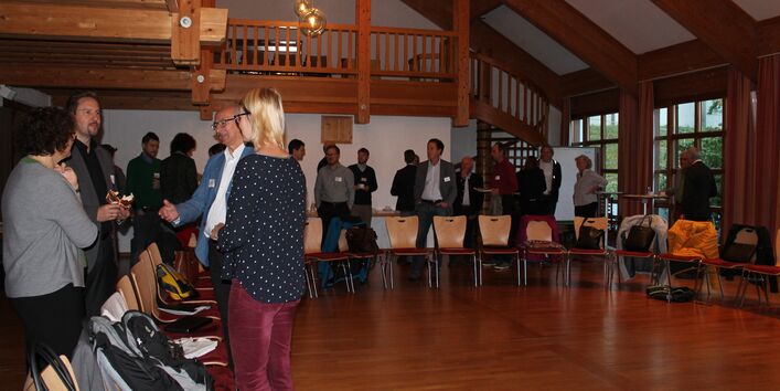 Teilnehmende stehen in gelockerter Atmosphäre im Veranstaltungsraum beim Stuhlkreis. Durch die großen Fenster geht der Blick ins Grüne.