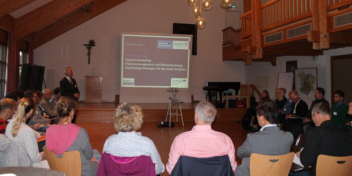 Teilnehmende sitzen im Stuhlkreis vor einer Präsentationswand und hören dem Vortragenden zu. Die Präsentationswand zeigt den Titel der Veranstaltung samt Logos, vorwiegend in blauen und weißen Farben.