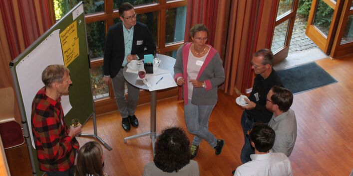 Teilnehmende stehen kreisförmig zusammen. Zwei Teilnehmende lehnen sich dabei auf einen Stehtisch und zwei weitere Teilnehmende halten Pausengetränke in ihren Händen. Im Hintergrund führt eine verglaste Tür nach draußen ins Grüne.