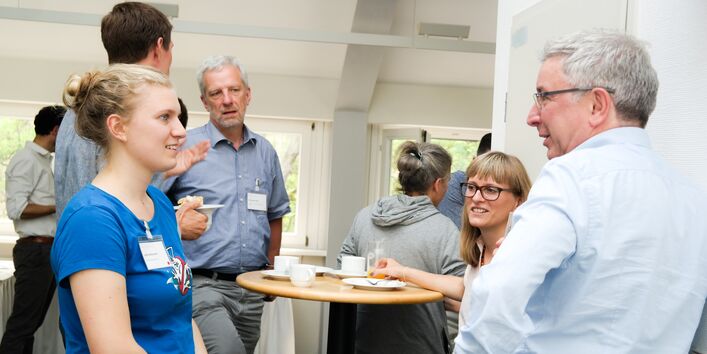 Kaffeepause während des Dialogs. Personen dabei stehen an Stehtischen und unterhalten sich. Der Raum ist in hellen Tönen gehalten. Im Vordergrund stehen eine Frau im blauen T-Shirt und ein Mann im hellfarbigen Hemd. Sie schauen sich an.