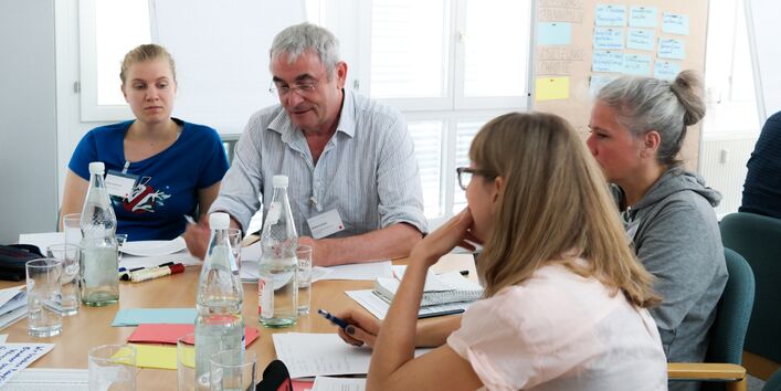 Planspiel: Teilnehmende sitzen an einem Gruppenarbeitstisch auf dem Getränke stehen und Papiere, farbige Moderationskarten liegen. Die Personen sehen nachdenklich aus.