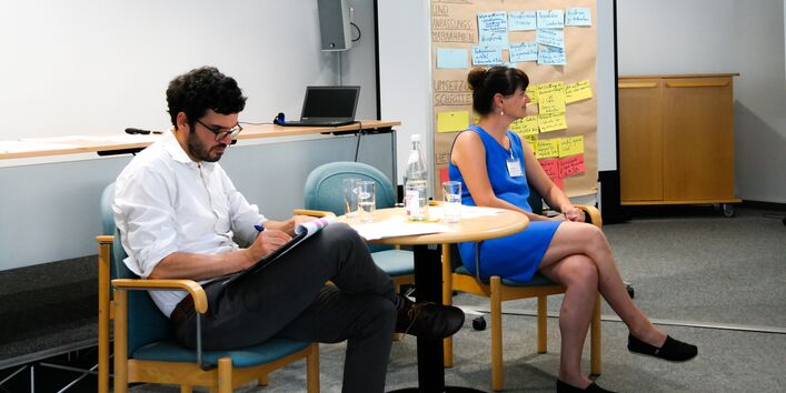 Reflexion: Die Durchführenden von der Veranstaltung sitzen zu zweit an einem Tisch. Sie schauen und hören in die Runde. Im Hintergrund steht eine Moderationswand, die mit diversfarbigen und beschrifteten Moderationskarten bestückt ist. Die Frau trägt ein blaues Sommerkleid. Der Mann ein helles Hemd und schwarze Hose.