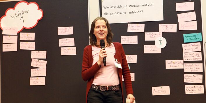 Teilnehmerin im rosa Shirt und roter Strickjacke erläutert die Ergebnisse aus dem Workshop: Wie lässt sich die Wirksamkeit von Klimaanpassung erhöhen? Sie spricht in ein Mikrofon, welches sie in der Hand hält.
