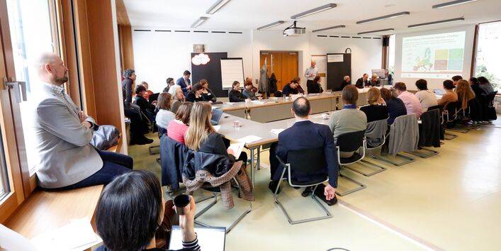 Workshopgruppenraum Ambiente im Umweltbundesamt: Teilnehmende sitzen im Rechteck an Tischen und hören dem Vortragenden zu. Der Raum ist in hellen Farben gestaltet. Manche Teilnehmende haben sich auf Stühlen und Fensterbänke verteilt.