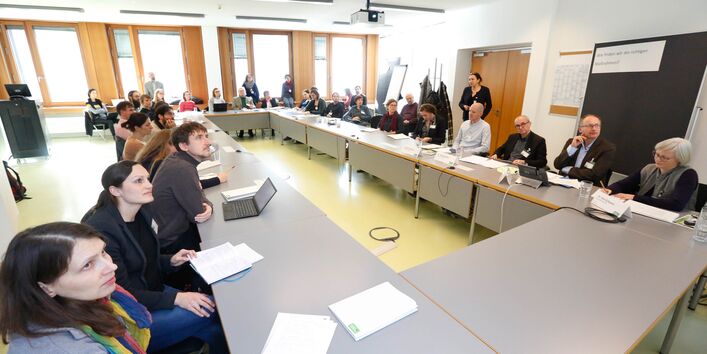 Teilnehmende sitzen an hellgrauen Tischen im Rechteck und schauen hoch zur Präsentationsfläche.