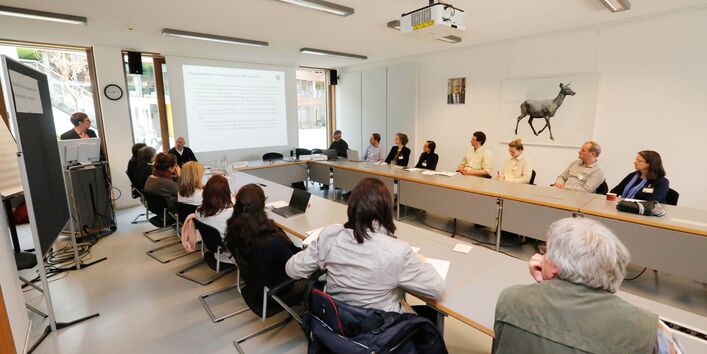 Teilnehmende sitzen an hellgrauen Tischen und schauen auf die Leinwand. Auf der Leinwand ist eine Präsentation zu sehen und im Hintergrund ist ein Bild mit einem großen Reh in Schwarz Weiss zu sehen. An der Decke hängt der Beamer.