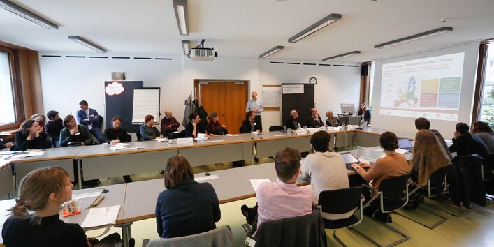 Teilnehmende sitzen an Tischen im Rechteck und folgen einer Präsentation. Die Präsentierenden stehen vorn am Stehpult und neben der Stellwand.