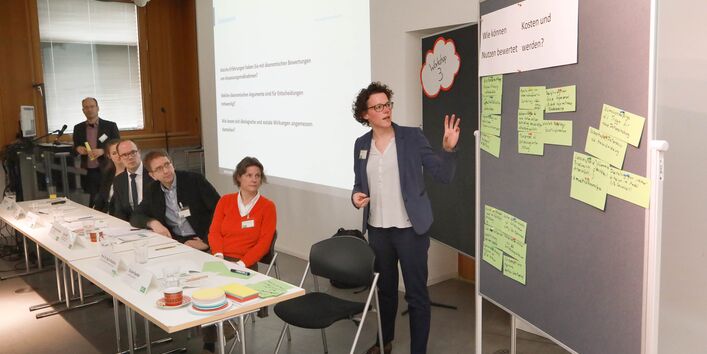 Eine Teilnehmerin präsentiert Gruppenarbeitsergebnisse an einer Pinwand. Die Ergebnisse stehen stehen auf hellgrünen Moderationskarten geschrieben. Im Hintergrund ist eine Präsentation eingeblendet. Davor steht ein Teilnehmender am Stehpult.