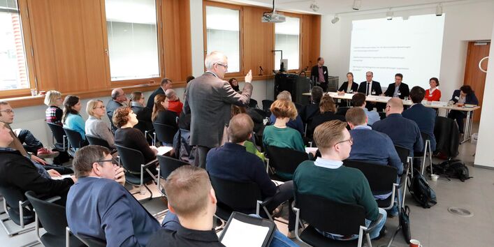 Ein Teilnehmer steht in mitten der Stuhlreihen vor dem Podium und spricht gestikulierend. Die anderen Teilnehmenden sitzen auf den Stühlen und hören nachdenklich zu.