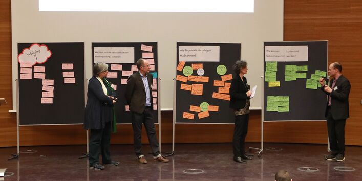 Abschlusspräsentation auf dem Podium im Hörsaal. Auf dem Podium stehen 4 Stellwände nebeneinander. Auf den Stellwänden befinden sich die Workshopergebnisse auf farbigen Moderationskarten geschrieben angepinnt. Und die jeweiligen Moderatorinnen und Moderatoren erläutern die Ergebnisse. Zwei Personen halten ein Mikrofon in der Hand.