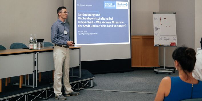 Vortragender steht neben einer Leinwand und hält den einführenden Vortrag. Auf der Leinwand ist das KomPass Log und der Veranstaltungstitel zu sehen. Der Votragende trägt eine helle Hose mit blauem Hemd.