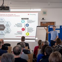 Eine Person referiert gestikulierend vor einem Flipchart. Im Vordergrund sind unscharf die Hinterköpfe des Publikums abgebildet. Im Hintergrund wird eine Präsentation zu projiziert.