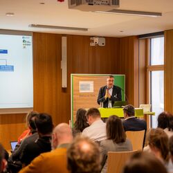 Eine Person referiert gestikulierend in einem Sitzungsraum. Im Vordergrund sind unscharf die Hinterköpfe des Publikums abgebildet. Im Hintergrund wird eine Präsentation projiziert.