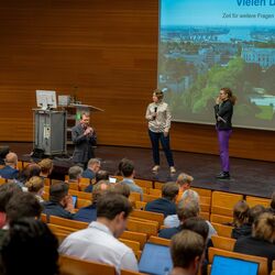 Eine Person spricht in der ersten Reihe des UBA-Hörsaals zum Publikum gewandt in ein Mikrofon. Zwei weitere Personen stehen zuhörend auf der Bühne. Im Vordergrund sind unscharf die Hinterköpfe des Publikums abgebildet. Im Hintergrund wird eine Präsentation projiziert.