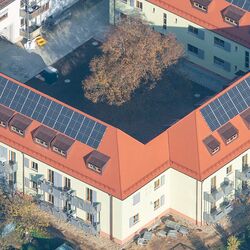 Luftbild eines Wohngebäudes mit drei Flügeln in einem U. Zwei Vollgeschosse und ein Walmdach mit mehreren Gauben und PV-Anlagen. Im Innenhof ein großer Baum, in der Umgebung Kleingärten. Vor der Öffnung des U ein linienförmiger Wohnbau ähnlicher Kubatur.