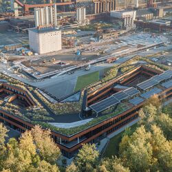 : Luftbild eines Industriegeländes. Im Vordergrund ein dreigeschossiger Bau mit einem L-Grundriss, Innenhöfen und einem Gründach. Im Hintergrund mehrere Schornsteine.