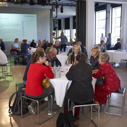 In einem großen Raum stehen mehrere runde Tischen, an denen diskutierende Menschen sitzen.