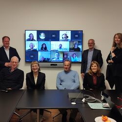 In a small meeting room, 7 people sit or stand in front of a screen. On the screen appear the faces of people in the meeting call.