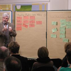 Ein Workshopteilnehmer stellt Workshopergebnisse, in Form von handgeschriebenen Karten die an Pinwänden befestigt sind, der gesamten Workshopgruppe vor.