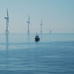 Das Foto zeigt drei Windenergieanlagen im Meer mit Jacket-Fundament (als Fachwerk konstruierte Gründungsstruktur). Ganz im Hintergrund ist die Forschungsplattform FINO1 sichtbar. Im Vordergrund ist ein im Verhältnis zu den Windenergieanlagen klein wirkendes Schiff sichtbar.