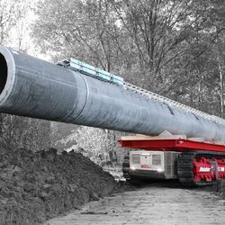 ein kleines selbstfahrendes Raupenfahrzeug transportiert auf seiner Ladefläche einen großen Betonmast durch den Wald