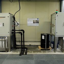 teschnische Anlage in einer Werkshalle aus zwei Boxen, aus denen Rohre in eine Gitterabdeckung im Fußboden führen