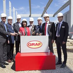 Sieben Männer und eine Frau haben sich anlässlich einer Grundsteinlegung mit Blauhelmen und Hämmern vor einem symbolischen Grundstein mit dem Firmenlogo "Graf" für ein Gruppenfoto aufgestellt 