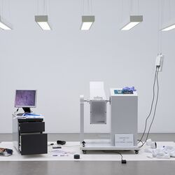 Kunstobjekt in einem Ausstellungsraum: Auf einem Rollcontainer steht ein PC-Monitor mit einem unscharfen Bild, daneben ein Gerät mit Schild "Laboratory of Electronic Ageing". Auf dem Boden liegen verstreut Ausdrucke, eine Rolle Klebeband und andere Objekte.