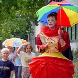 Eine Frau als Clown verkleidet hält einen bunten Regenschirm und läuft mit einigen Kindern auf einem Bürgersteig.