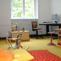Eltern-Kind-Zimmer: Am Fenster steht ein Schreibtisch umgeben von Spielsachen, einer Spielküche, einer Sitzecke, im Vordergrund eine Matratze mit großem Kissen.