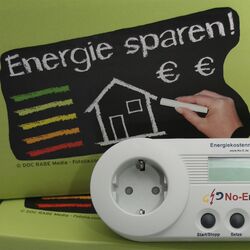 Das Bild zeigt eine hellgrüne Pappkiste mit einem Messgerät für Leerlaufverluste von Elektrogeräten im Vordergrund