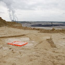 Eine karge Bergbau Landschaft, im Hintergrund eine Energieanlage, vorne sieht man den roten Rahmen, mit dem die Künstlerin einen Bodenabdruck macht.