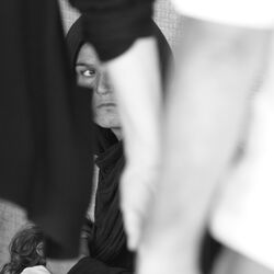 Eine Frau sitzt mit ihrem Kind im Arm am Straßenrand, zwei Passanten gehen an ihnen vorbei.