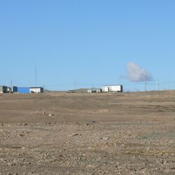 Im Hintergrund stehen einige Hallen und Häuser, vorne ist die karge Landschaft der Arktis zu sehen. 