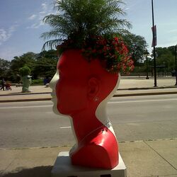 Auf dem Bürgersteig steht ein übergroßer roter Kopf, er dient als Blumentopf.