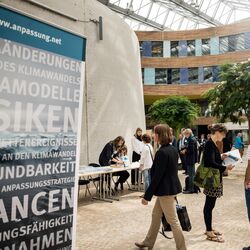 Sie sehen ein Bild vom Nationaler Dialog 2017 Gemeinsam sind wir klimaresilient – Wie Beteiligung in Städten und Regionen gelingt. Das Bild wurde im Umweltbundesamt in Dessau aufgenommen. Auf der rechten Seite sehen Sie ein Aufsteller zum Thema Klimarisiken und Anpassung. Auf der rechten Seite des Bildes sind Menschen auf dem Weg in den Hörsaal in Dessau.
